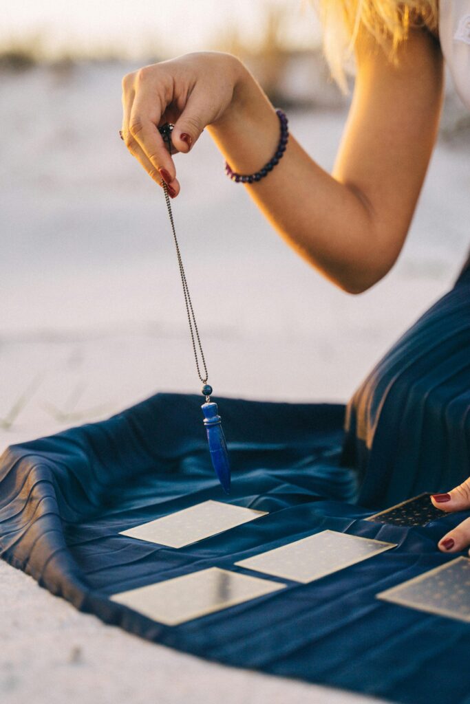A woman's hand holding a pendulum over tarot cards on a beach at sunset.